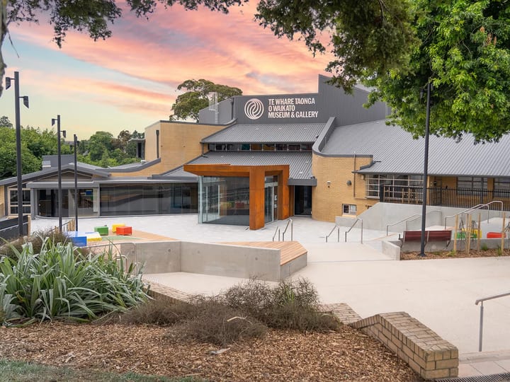 Brick and metal-roof building with timber-framed glazed entry, concrete forecourt, gardens at dusk