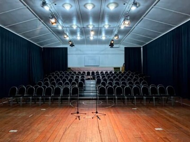 Hall interior with rows of chairs facing a draped stage lit in blue and purple