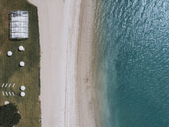 Aerial view of glass marquee and white umbrellas on grass lawn beside white sand beach and blue water