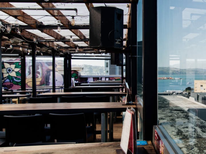 Open rooftop bar interior by day with exposed steel beams, long wooden benches, and large window overlooking city