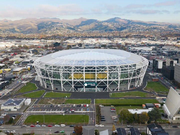 Large oval stadium with white lattice steel frame and translucent roof, aerial view in an urban setting with hills behind