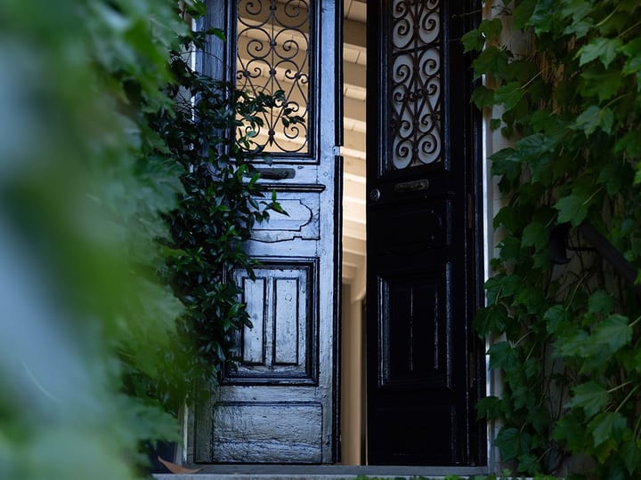 Dark iron-wrought entrance doors with ornate scrollwork panels, ajar to reveal lit interior, stone steps and climbing plants
