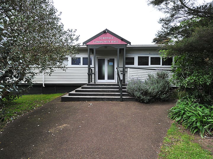 White weatherboard hall with entry steps and pink fascia sign, surrounded by planting