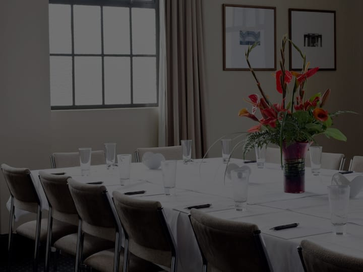 Boardroom with long white-clothed table, upholstered chairs, two framed prints, tall windows