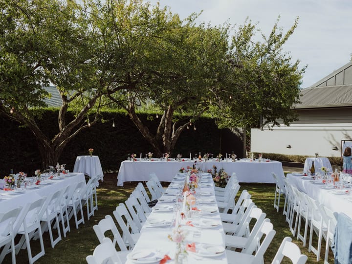 Outdoor reception with white linen tables, white folding chairs, festoon lights and mature trees