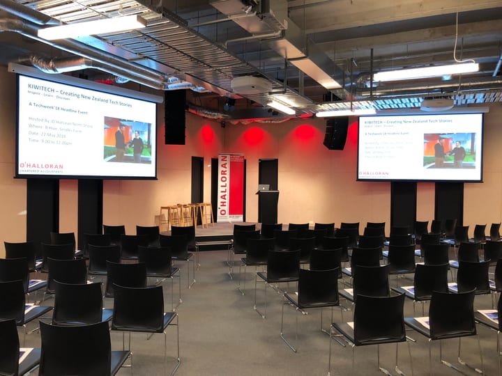 Event hall set up in theatre style with rows of black chairs, dual screens, and red uplighting