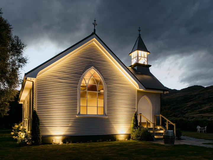 White timber church exterior lit at night with glowing arched window and dark hill beyond