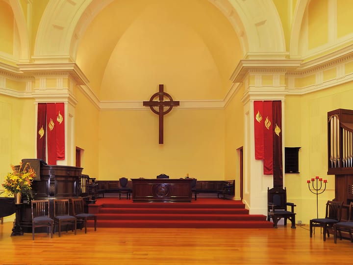 Pale yellow church sanctuary with arched apse, wooden cross, pipe organ, and red carpet aisle