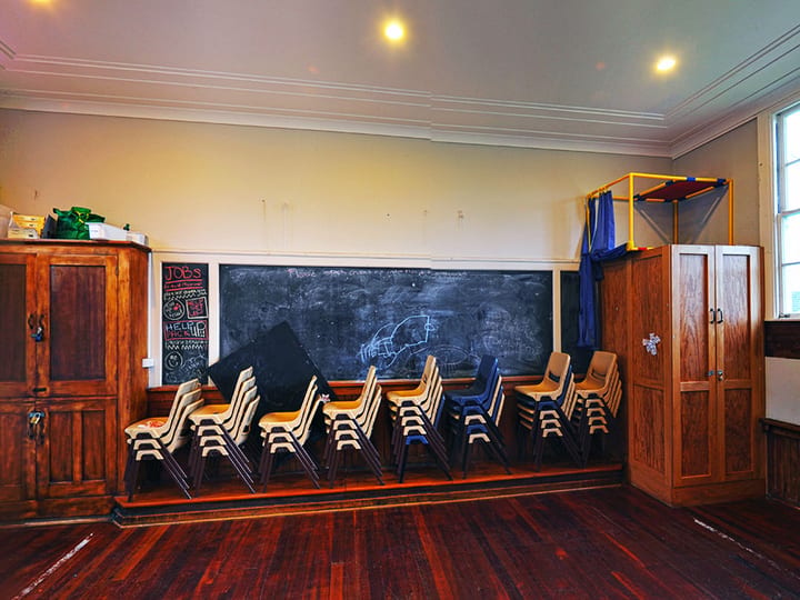 Plain hall interior with stacked school chairs, timber floor, chalkboard, and wooden storage cupboards