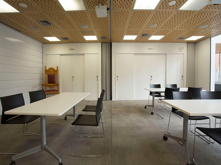 Two adjoining meeting rooms with perforated timber ceiling, movable white tables and dark chairs on carpet