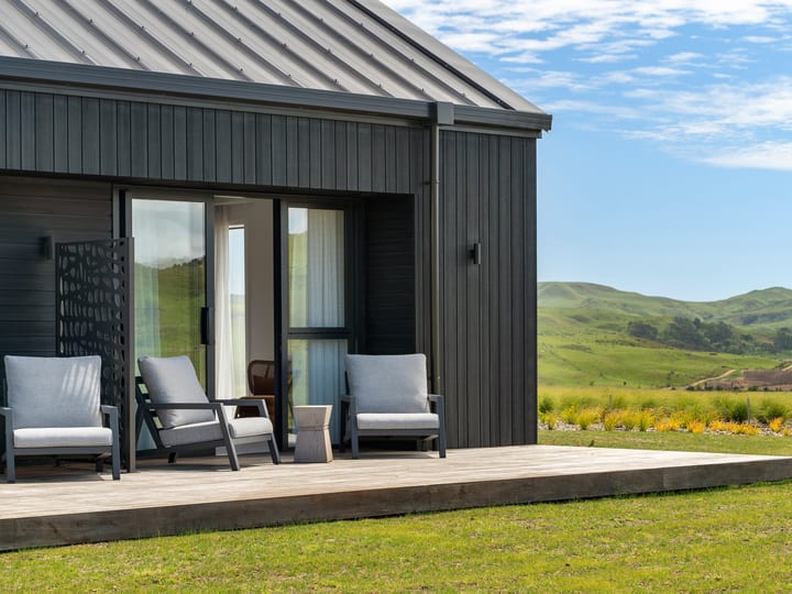 Dark vertical-clad building with timber deck, two outdoor chairs, and green hillside view