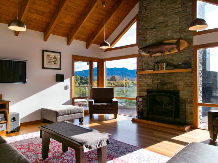 Open-plan lounge with exposed timber beams, stone fireplace, and floor-to-ceiling windows to mountains