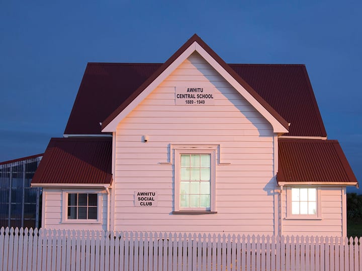 White weatherboard hall with red iron roof and picket fence, lit at dusk