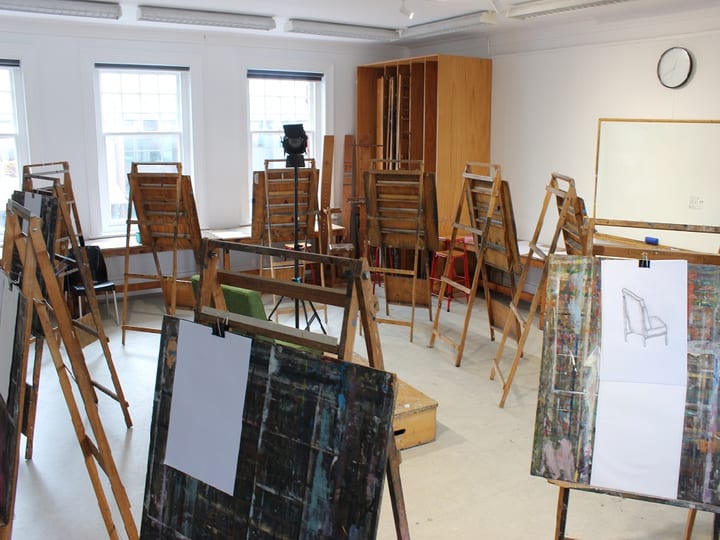Bright studio room with multiple timber easels and canvases, whiteboard, and tall windows
