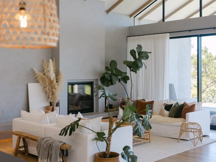 Open living room with timber beams, concrete fireplace, white sofas, indoor plants, and glass walls