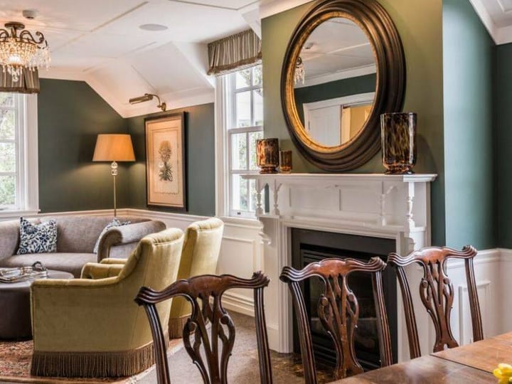 Green-walled lounge with white fireplace, chandelier, timber dining chairs and velvet armchairs