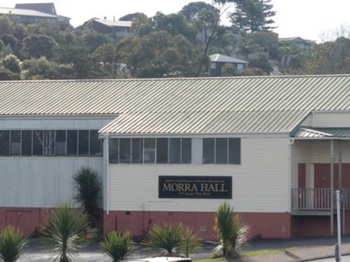Low-pitched cream and red-brick community hall exterior with green corrugated roof, hillside behind