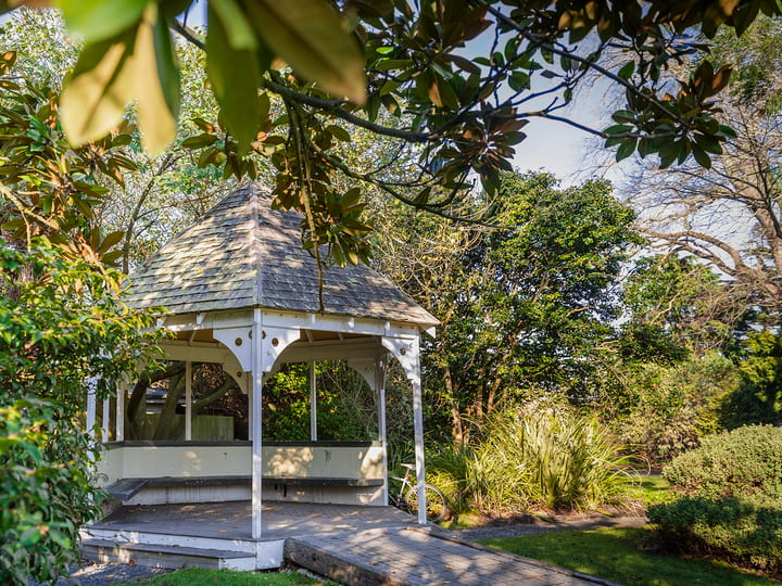 White wooden gazebo with shingled roof set among dense mature trees and garden
