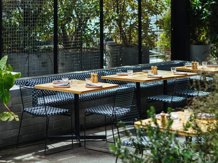 Outdoor dining terrace with timber tables, black wire chairs, and dense green plantings around black steel screen