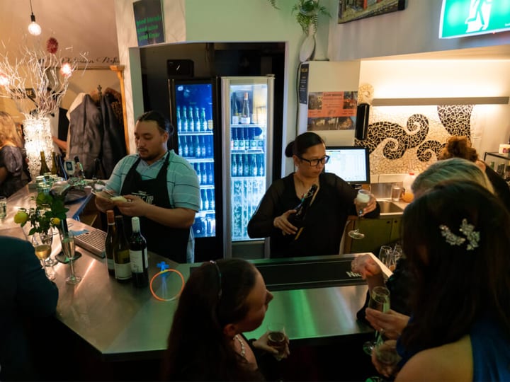 Bar service area with illuminated drinks fridge and bartender, decorated interior space with guests in background