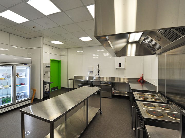Commercial kitchen with stainless benches, extraction hood, range cooker, glass-front fridge, and dark floor