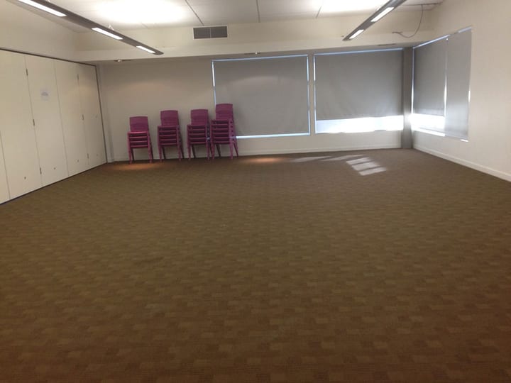 Empty carpeted hall with stacked pink chairs and roller blinds on windows