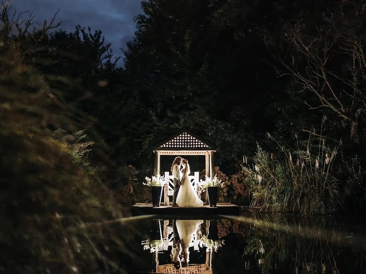 Illuminated gazebo on a jetty reflected in a still pond at night, surrounded by trees