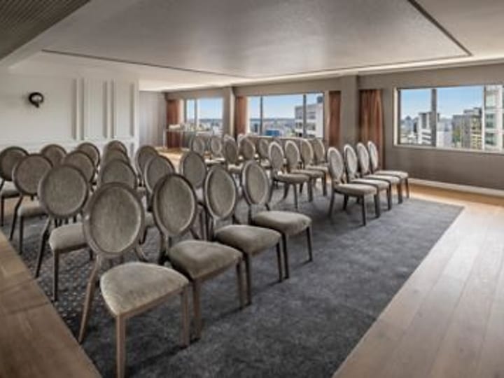 Neutral-toned meeting room with rows of upholstered chairs, two large windows, pale timber flooring