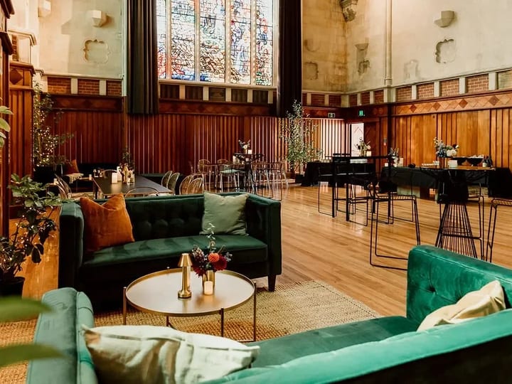 Church hall interior with dark timber panelling, stained-glass windows, and velvet sofas
