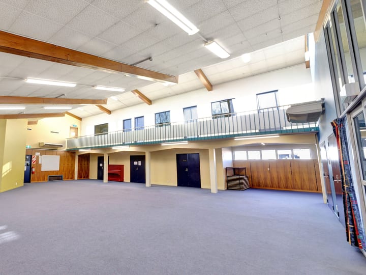 Large cleared hall with grey carpet, exposed timber roof beams, mezzanine balcony, and high windows