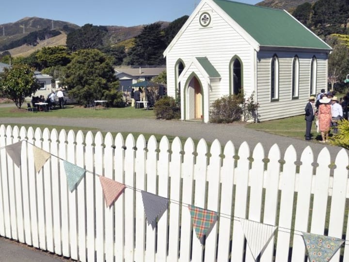 Small white timber chapel with green roof and arched entry, beside a white picket fence with bunting
