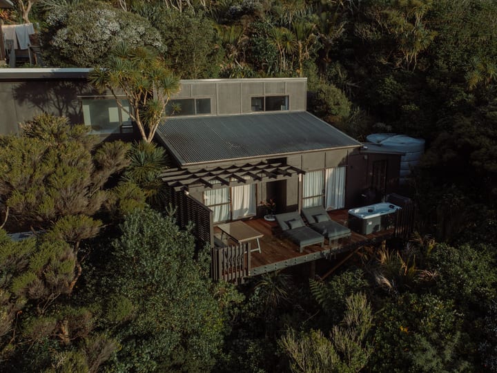 Corrugated roof cabin with timber deck, spa bath and lounge chairs surrounded by bush, aerial view