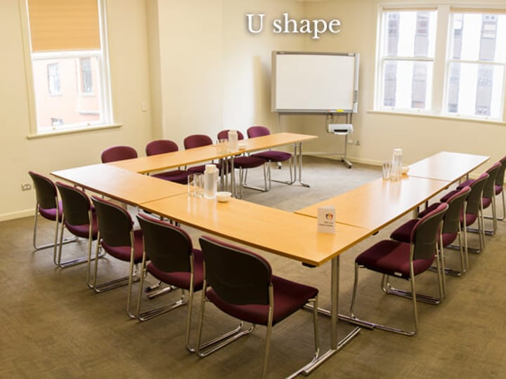 Carpeted meeting room with U-shaped timber tables, purple chairs, and whiteboard by windows