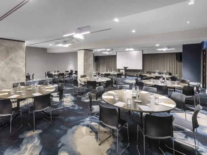 Hotel conference room with round tables, chairs, projector screen, and patterned carpet