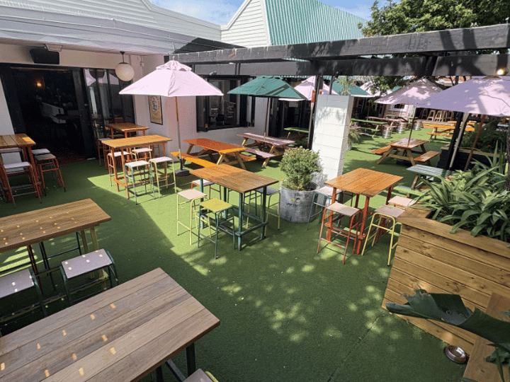 Open-air courtyard with timber picnic tables, coloured stools, market umbrellas, and turf underfoot