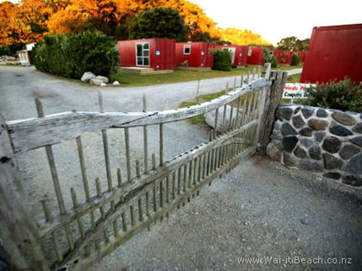 Rustic wooden entrance gate onto a gravel driveway with red cabin buildings beyond at dusk