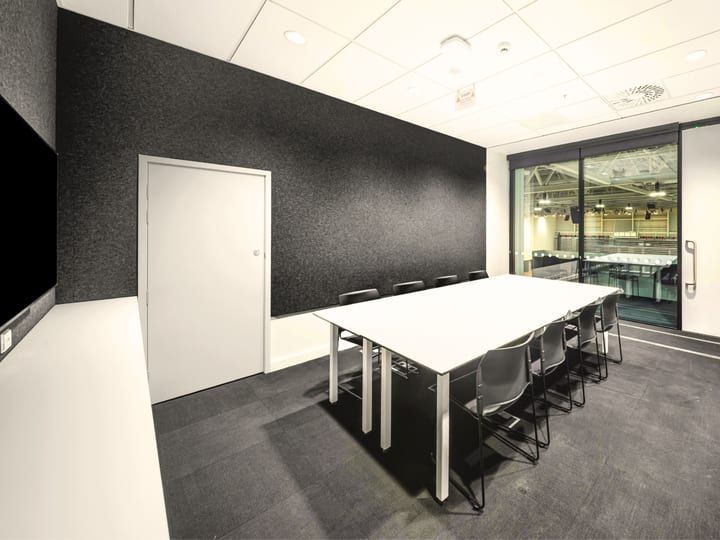 Small meeting room with round-ended white table, grey chairs, wall screen and dark acoustic walls