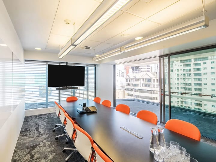 Long meeting room with dark table, orange chairs, wall-mounted screen, and city-view windows on two sides