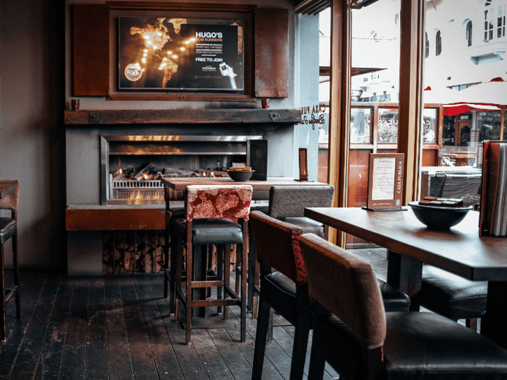 Dim bar room with open fireplace, upholstered high stools, dark timber tables, and street-facing windows