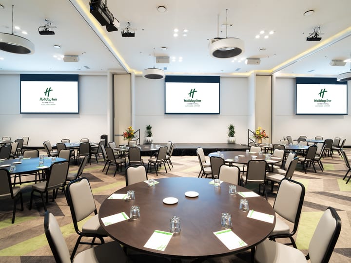 Large carpeted function room with round tables, three projection screens, and recessed ceiling lighting