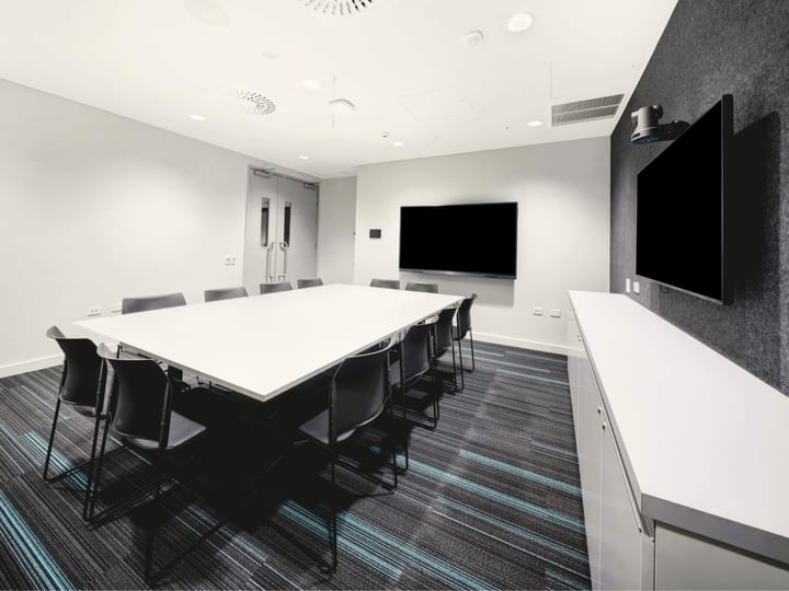 White meeting room with long white table, black chairs, two wall-mounted screens and patterned carpet
