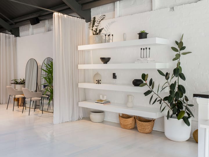 Bright white studio with open shelving, potted plants, sheer curtain partition, and arched mirrors