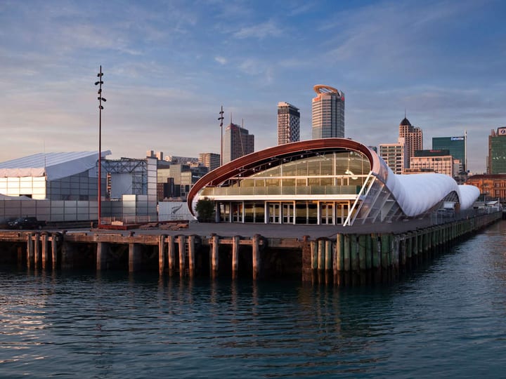White barrel-vault pavilion on pier over water, Auckland city skyline at dusk behind