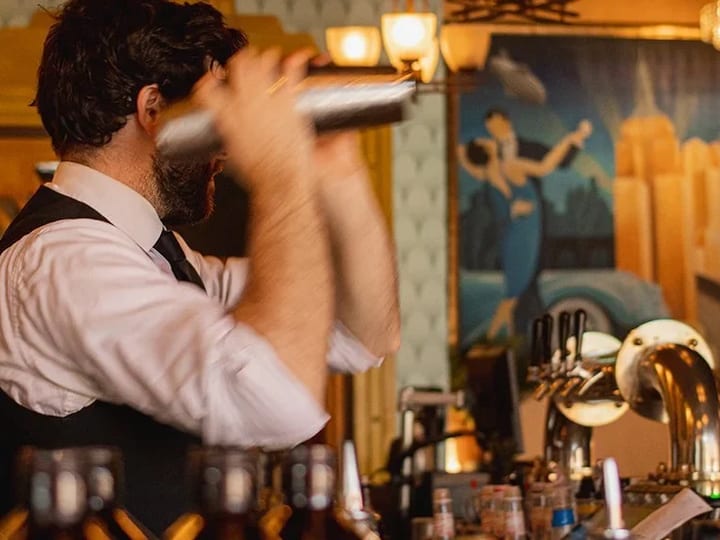 Bartender shaking cocktail behind bar with art deco mural and beer taps