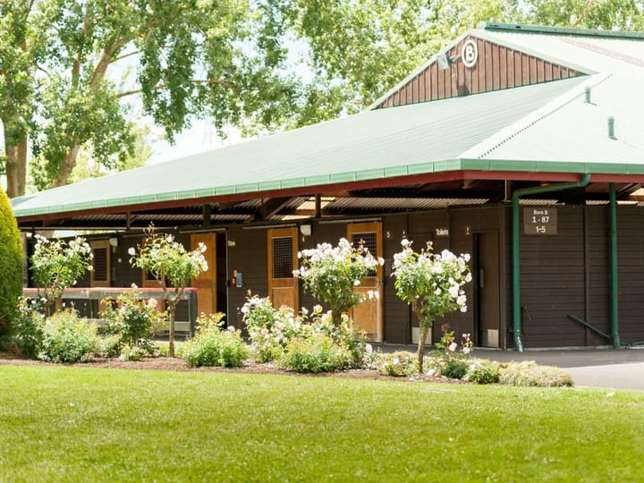 Dark timber stable block with green roof, flowering plants along the front, and lawn in foreground