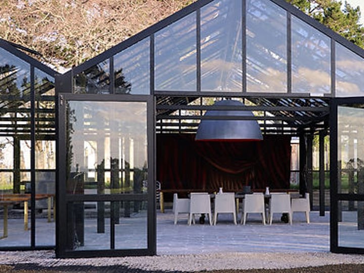 Black steel glasshouse interior with white chairs and dark hanging pendant, surrounded by trees