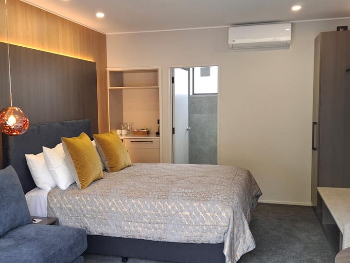 Hotel room with large double bed, yellow cushions, timber-panel feature wall, pendant light, and ensuite beyond