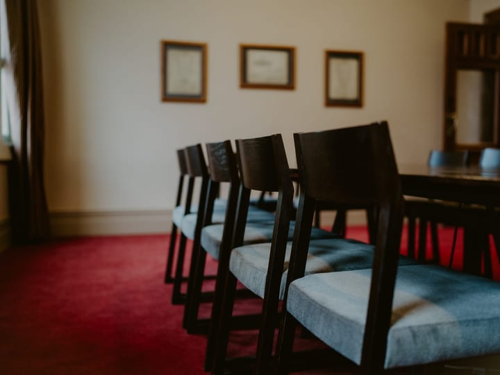 Dark-toned room with dark wood chairs and blue upholstered seats around a long table, framed prints on wall