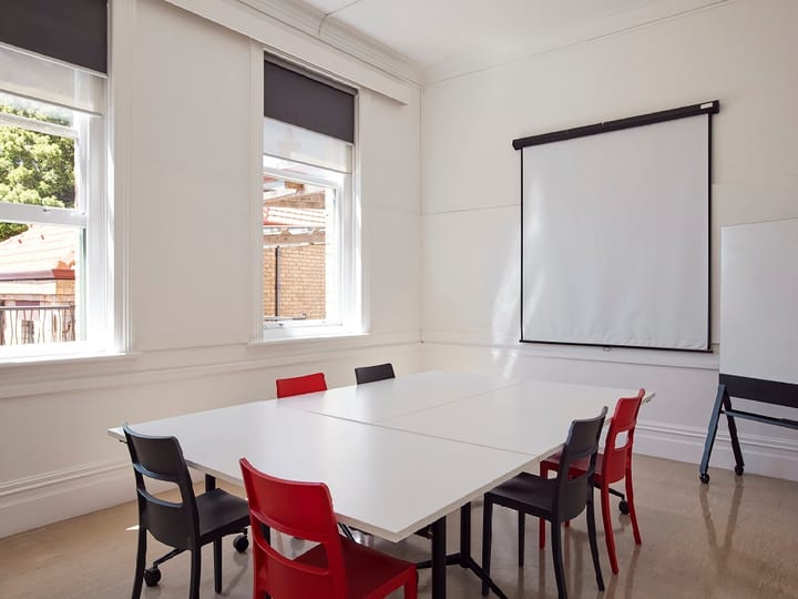 Small white room with table, red and dark chairs, projection screen, and two sash windows