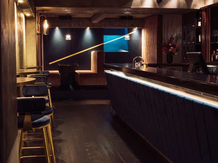 Dark intimate bar with navy velvet stools, lit bar counter, and pink booth seating